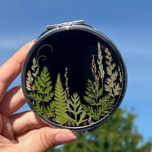May include: A round, silver compact mirror with a black background. The mirror features a botanical design of green ferns and grasses. The compact is held against a bright blue sky.