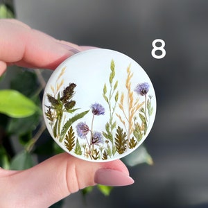 Pode incluir: Um suporte de telefone redondo branco com um design bot&acirc;nico. O design apresenta flores e gram&iacute;neas secas em tons de roxo, verde e dourado. O n&uacute;mero "8" &eacute; impresso em branco no lado direito do suporte.