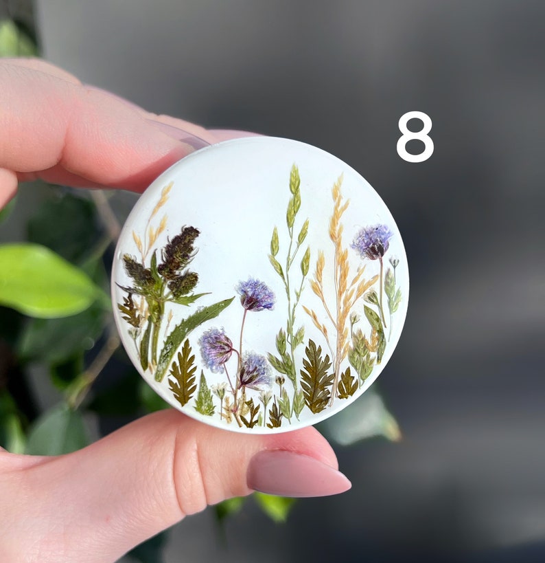 Pode incluir: Um suporte de telefone redondo branco com um design bot&acirc;nico. O design apresenta flores e gram&iacute;neas secas em tons de roxo, verde e dourado. O n&uacute;mero "8" &eacute; impresso em branco no lado direito do suporte.