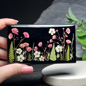 May include: A black rectangular resin tray with a pressed flower design. The design features pink and white flowers and green leaves on a black background.
