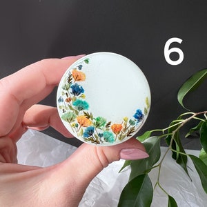 Pode incluir: Um objeto decorativo redondo e branco com um design floral em tons de azul, laranja e verde. O n&uacute;mero "6" &eacute; vis&iacute;vel. O objeto &eacute; segurado em uma m&atilde;o.