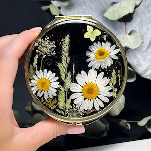 May include: A gold compact mirror with a black background and pressed white daisies and greenery.