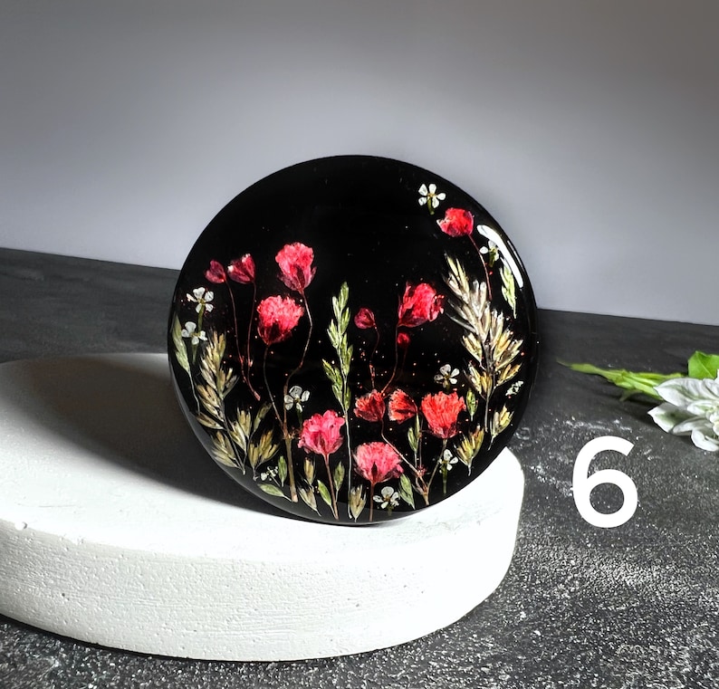 K&ouml;nnte beinhalten: Ein runder schwarzer Untersetzer mit einem Blumendesign aus roten und wei&szlig;en gepressten Blumen. Der Untersetzer befindet sich auf einer wei&szlig;en Oberfl&auml;che.