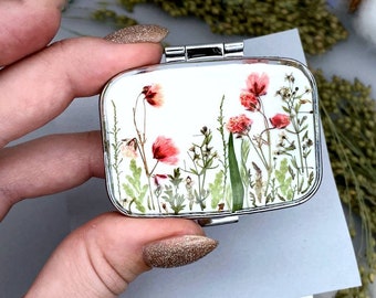 Op de afbeelding: Een zilveren compact spiegel met een witte achtergrond en geperste bloemen erin. De bloemen zijn roze en wit en hebben groene stelen en bladeren.
