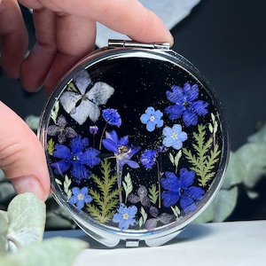 May include: A round, silver compact mirror with a black background. The mirror's face features an arrangement of dried blue and white flowers and green foliage. The compact is held open, revealing the mirror's reflective surface.