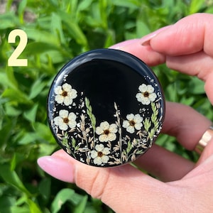 K&ouml;nnte beinhalten: Ein schwarzer Handy-Griff aus Harz mit wei&szlig;en gepressten Blumen und gr&uuml;nem Laub. Der Griff wird von einer Hand gehalten.