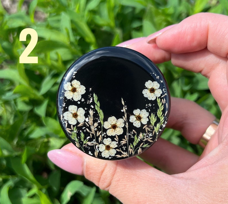 K&ouml;nnte beinhalten: Ein schwarzer Handy-Griff aus Harz mit wei&szlig;en gepressten Blumen und gr&uuml;nem Laub. Der Griff wird von einer Hand gehalten.