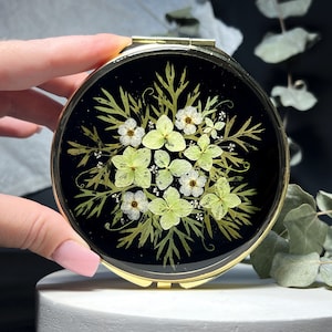May include: A round gold-toned compact mirror featuring a black surface decorated with pressed light green hydrangea petals, small white flowers, and delicate green fern-like foliage arranged in a circular botanical pattern on the front cover.