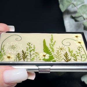 Op de afbeelding: Een zilveren metalen visitekaartjeshouder met een cr&egrave;mekleurige achtergrond met een ontwerp van geperste bloemen. Het ontwerp bevat witte bloemen, groene bladeren en varens.