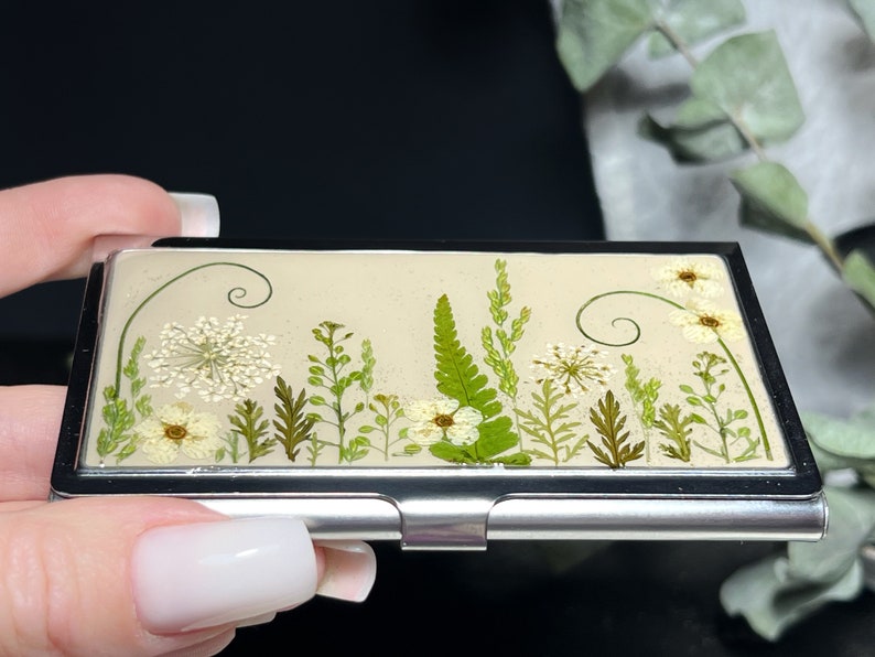 Op de afbeelding: Een zilveren metalen visitekaartjeshouder met een cr&egrave;mekleurige achtergrond met een ontwerp van geperste bloemen. Het ontwerp bevat witte bloemen, groene bladeren en varens.