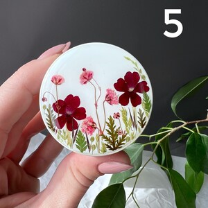 Pode incluir: Um suporte de telefone redondo em resina branca com um design floral. O design inclui flores vermelhas escuras e rosa, folhas verdes e caules delicados. O n&uacute;mero "5" &eacute; vis&iacute;vel.