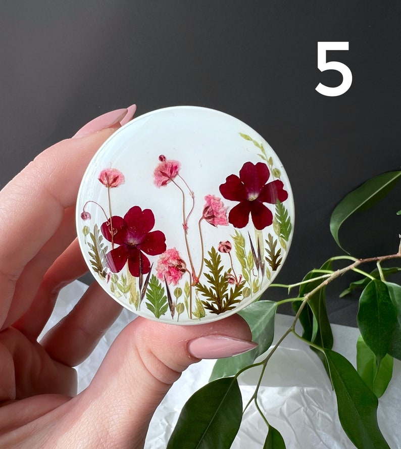 Pode incluir: Um suporte de telefone redondo em resina branca com um design floral. O design inclui flores vermelhas escuras e rosa, folhas verdes e caules delicados. O n&uacute;mero "5" &eacute; vis&iacute;vel.