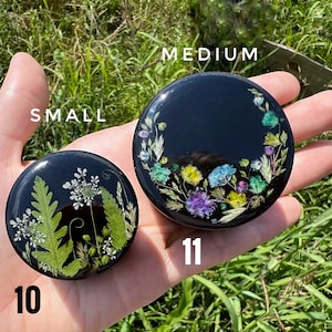 K&ouml;nnte beinhalten: Zwei runde, schwarze Harzkistchen in verschiedenen Gr&ouml;&szlig;en mit Blumendesigns. Die kleinere Box hat ein gr&uuml;nes Blumendesign und ist mit "10" beschriftet. Die gr&ouml;&szlig;ere Box hat ein farbenfrohes Blumendesign und ist mit "11" beschriftet. Der Text "SMALL" und "MEDIUM" befindet sich &uuml;ber den Boxen.