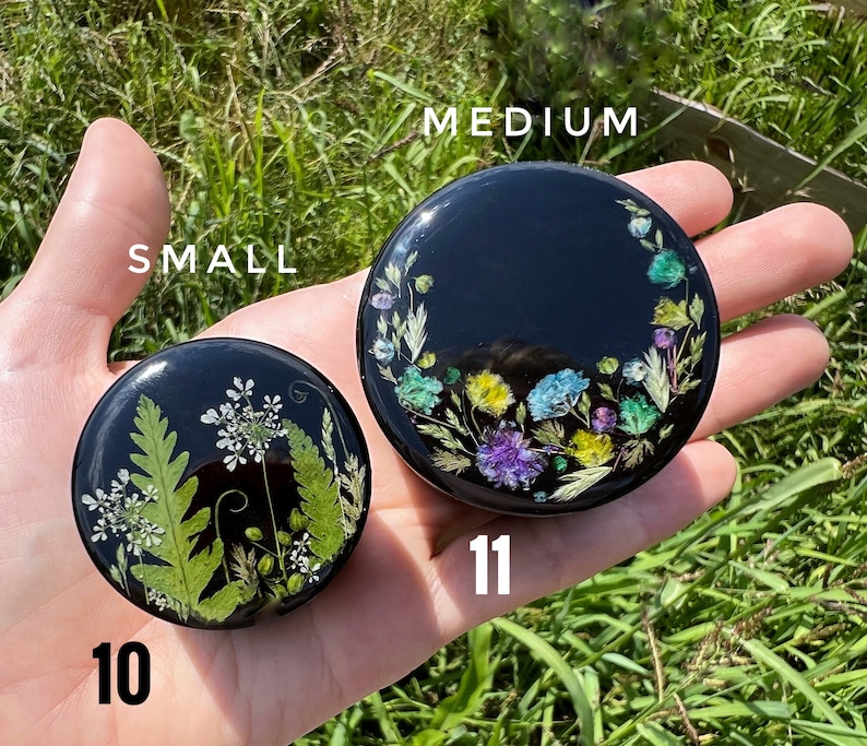K&ouml;nnte beinhalten: Zwei runde, schwarze Harzkistchen in verschiedenen Gr&ouml;&szlig;en mit Blumendesigns. Die kleinere Box hat ein gr&uuml;nes Blumendesign und ist mit "10" beschriftet. Die gr&ouml;&szlig;ere Box hat ein farbenfrohes Blumendesign und ist mit "11" beschriftet. Der Text "SMALL" und "MEDIUM" befindet sich &uuml;ber den Boxen.