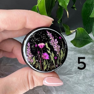 K&ouml;nnte beinhalten: Ein schwarzer Handy-Griff aus Harz mit einer runden, klaren Epoxidkuppel, die gepresste lila und rosa Blumen und gr&uuml;ne Bl&auml;tter auf einem schwarzen Hintergrund mit goldenem Glitzer zeigt.