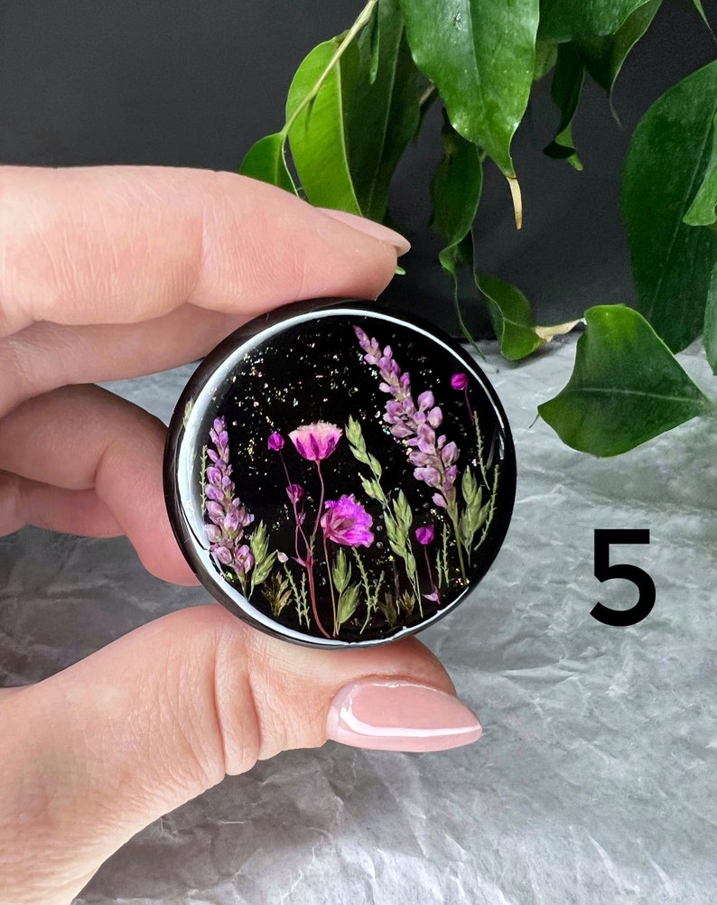 K&ouml;nnte beinhalten: Ein schwarzer Handy-Griff aus Harz mit einer runden, klaren Epoxidkuppel, die gepresste lila und rosa Blumen und gr&uuml;ne Bl&auml;tter auf einem schwarzen Hintergrund mit goldenem Glitzer zeigt.