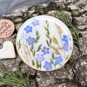 Pode incluir: Uma base de resina redonda com borda dourada, apresentando delicados mios&oacute;tis azuis, grama verde e trigo seco. A base est&aacute; sobre um fundo de madeira natural, mostrando uma est&eacute;tica artesanal.