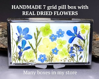 Peut inclure: Une boîte à pilules en argent avec un fond blanc et un motif de fleurs séchées pressées dans des tons de bleu, jaune et vert. Le texte "HANDMADE 7 grid pill box with REAL DRIED FLOWERS" est au-dessus de la boîte et le texte "Many boxes in my store" est en dessous de la boîte.