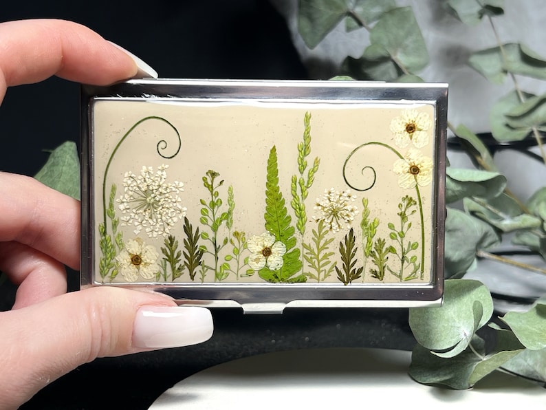 Op de afbeelding: Een zilveren kaarthouder met een ontwerp van geperste bloemen. Het ontwerp bevat witte bloemen, groene bladeren en stelen op een cr&egrave;mekleurige achtergrond.