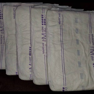 May include: Several white adult diapers are stacked, each with purple accents and text. The diapers are labeled "MoliCare Premium Slip M super plus". The image is taken from a slightly elevated angle, showing the products in a neat arrangement.