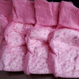 May include: A stack of pink diapers with a heart pattern. The diapers are folded and stacked, with a ruffled edge at the top. The background is dark.