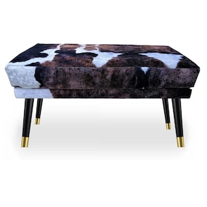 Könnte beinhalten: Ein rechteckiger Hocker mit einem Kuhfellmuster in Braun, Schwarz und Weiß. Er hat vier schwarze Beine mit goldfarbenen Spitzen. Das Design ist modern und das Möbelstück wird wahrscheinlich zum Sitzen oder als Fußstütze verwendet. Maße in Zentimetern.