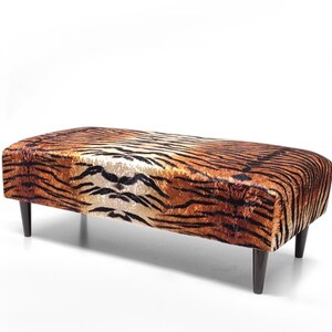 Peut inclure: Un pouf rectangulaire avec un motif de rayures de tigre dans les tons marron et noir. Le dessus rembourré repose sur quatre pieds effilés brun foncé. Le design évoque un imprimé animalier.