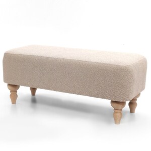 May include: A rectangular bench with a light beige, textured fabric covering. The bench has rounded corners and is supported by four turned, light-colored wooden legs. The overall design is simple and elegant, suitable for various interior styles.