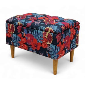 Op de afbeelding: Een zwarte opbergottoman met een bloemenprint met rode hibiscusbloemen, groene bladeren en granaatappels. De ottoman heeft een gestoffeerde bovenkant en houten poten.