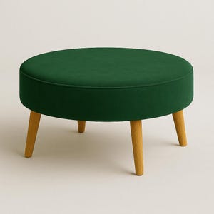 Può includere: Un pouf rotondo in velluto verde smeraldo con base in legno chiaro. La seduta bassa è sostenuta da quattro gambe affusolate e angolate. Il cuscino della seduta è di colore verde scuro uniforme. Il pouf ha un diametro di circa 76 cm.