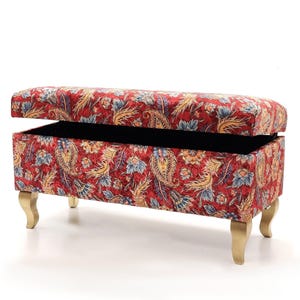 May include: A red storage bench with a floral pattern in blue, yellow, and gold. The bench has a hinged lid, revealing a black interior. It stands on four gold-coloured, curved legs, offering both style and functionality.