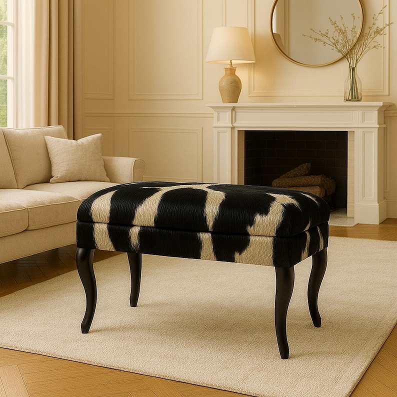 May include: A rectangular ottoman with a black and white cowhide pattern. The ottoman has four curved, dark-stained wooden legs. The piece is in a living room setting with a sofa and fireplace.