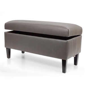 May include: A brown faux leather storage bench with a hinged top that opens to reveal storage space. The bench has four black wooden legs.