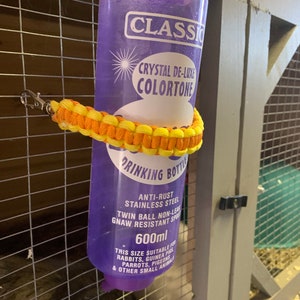 May include: Purple plastic drinking bottle with a yellow and orange braided strap. The bottle has a label that reads "Classic Crystal De-Luxe Colortone Drinking Bottle" and "Anti-Rust Stainless Steel Twin Ball Non-Leak Gnaw Resistant Spout 600ml". The label also states "This size suitable for Rabbits, Guinea Pigs, Parrots, Pigeons & other small animals."