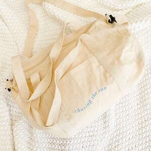 Puede incluir: Una bolsa de lona beige con cierre de cremallera y dos correas ajustables. La bolsa tiene un bolsillo grande en la parte delantera y el texto "chasing the sun" bordado en azul en el lateral.