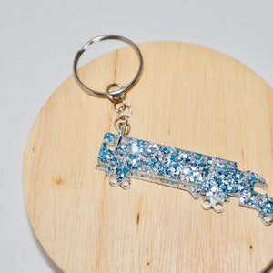 Peut inclure: Un porte-clés en forme de camion recouvert de paillettes argentées et bleues. Le porte-clés est attaché à un anneau de clé argenté.