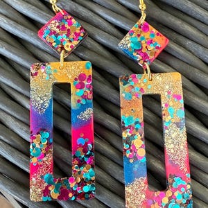 May include: A pair of vibrant dangle earrings with gold-coloured hooks. Each earring has a diamond-shaped top and a rectangular bottom with a cutout. The earrings are filled with glitter in shades of pink, blue, gold, and teal.