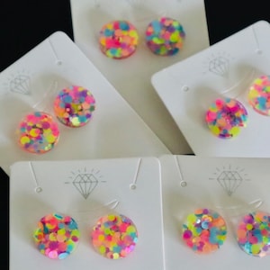 May include: Six pairs of colorful confetti stud earrings displayed on white card stock with a diamond logo. The earrings are made of clear resin with embedded confetti in pink, yellow, blue, and green.