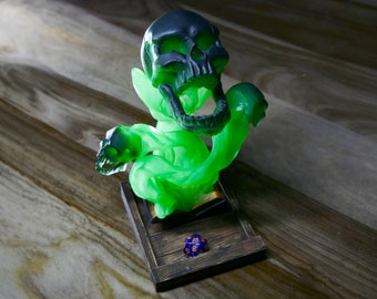 May include: A green and black skull figurine with a green, swirling, smoke-like base. A blue and gold twenty-sided die sits on a wooden base in front of the figurine.