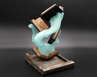 May include: A fantasy sculpture depicting open books suspended in a swirling, pale blue form. The books have wooden covers and pages, with one showing an engraved symbol. The base is a wooden tray, giving a rustic feel.