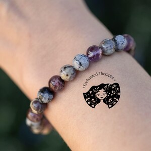 May include: A bracelet made of gray, white, and purple stones. The bracelet is on a person's wrist. The text "Enchanted Therapist." is on the person's arm.