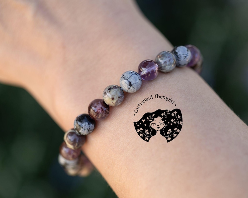 May include: A bracelet made of gray, white, and purple stones. The bracelet is on a person's wrist. The text "Enchanted Therapist." is on the person's arm.