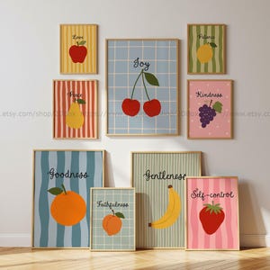 May include: A collection of nine framed wall art prints featuring various fruits and inspirational words. The prints display a red apple labeled Love, a yellow pear labeled Peace, an orange labeled Goodness, a peach labeled Faithfulness, a banana labeled Gentleness, a strawberry labeled Self-control, a bunch of grapes labeled Kindness, a lemon labeled Patience, and cherries labeled Joy.