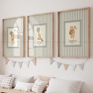 May include: Three framed Winnie the Pooh prints with a light wood frame. Each print features a different illustration of Pooh Bear with inspirational text. The background has vertical stripes in cream and light blue. A garland of small flags hangs below the prints.