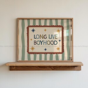 Pode incluir: Uma impressão artística emoldurada com o texto LONG LIVE BOYHOOD em letras azuis e castanhas. O design inclui uma borda recortada castanha, estrelas douradas e azuis, sobre um fundo de riscas verticais verde-sálvia e creme.