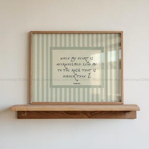 May include: A framed wall art print featuring a light sage green and white vertical striped background. The centre contains a rectangular cream-coloured panel with the black text: When my heart is overwhelmed: lead me to the rock that is higher than I. Psalm 61:2.