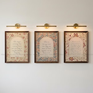 Könnte beinhalten: Drei rechteckige Wandbilder mit dunkelbraunen Holzrahmen, die jeweils mit einer messingfarbenen Leuchte versehen sind. Die Drucke zeigen florale Muster, die zentrale Textfelder mit handschriftlicher Kalligrafie und religiösen Versen umrahmen.
