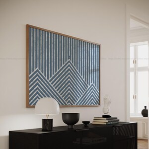 May include: A framed abstract print with a blue and white geometric pattern hangs on a white wall above a black console table. A white lamp with a black base sits on the table, along with a black bowl and a small white sculpture.