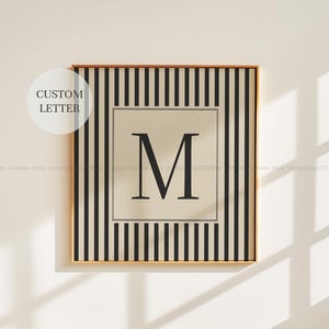 May include: A framed print with a large black letter "M" on a beige background, surrounded by black and white vertical stripes. The words "CUSTOM LETTER" are in a white circle in the upper left corner.