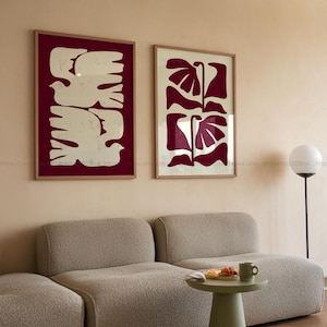 Può includere: Due stampe artistiche incorniciate. Una presenta un disegno di colomba color crema su sfondo bordeaux. L'altra stampa mostra un disegno floreale bordeaux su sfondo crema. Entrambe sono in cornici di legno e sono appese a una parete beige.